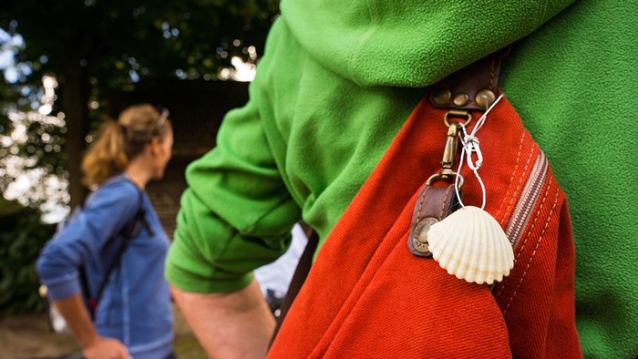 Zwei Fahrradfahrer*innen. Von der Person im Vordergrund sieht man nur den Rücken und einen Rucksack mit einem Muschelanhänger - dem Symbol des Jakobswegs.