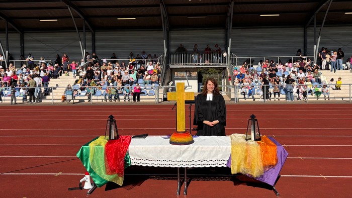Im Hintergrund: Die Tribüne des Stadions, die sich vor dem Gottesdienst füllt. Im Vordergrund: Der Altar mit einem bunten Kreuz und bunten Tüchern. Hinter dem Altar steht Pfarrerin Annika Wilinski im Talar.