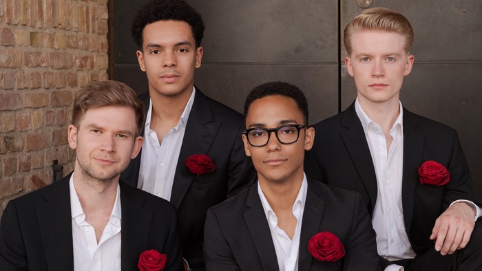 Vier Männer in formeller Kleidung mit roten Boutonnières posieren gemeinsam drinnen.