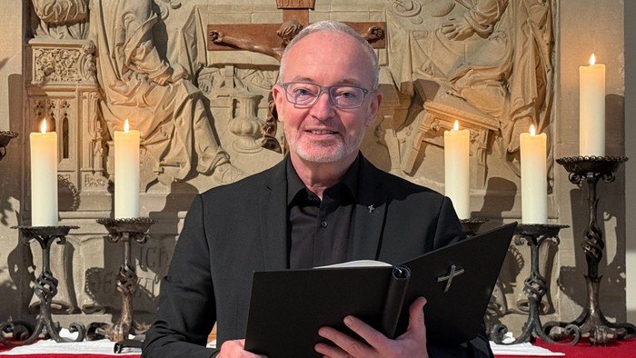 Ein Mann in formeller Kleidung hält Bücher in einer von Kerzenlicht erleuchteten Kirche und lächelt zum Altar.