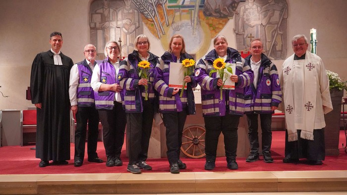Drei Personen in violetter Einsatzkleidung stehen, gesäumt von weiteren Beteiligten, mit Urkunden und Sonnenblumen vor dem Altar in einer Kirche.