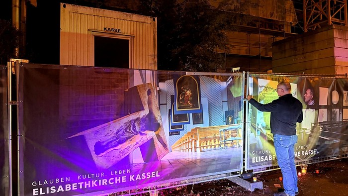 Ein Mann richtet nachts ein großes beleuchtetes Werbebanner für die Elisabethkirche Kassel aus.