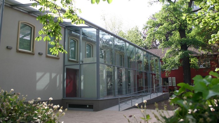 Modernes Gebäude mit großer Glaserweiterung, umgeben von üppigem Grün und Bäumen.