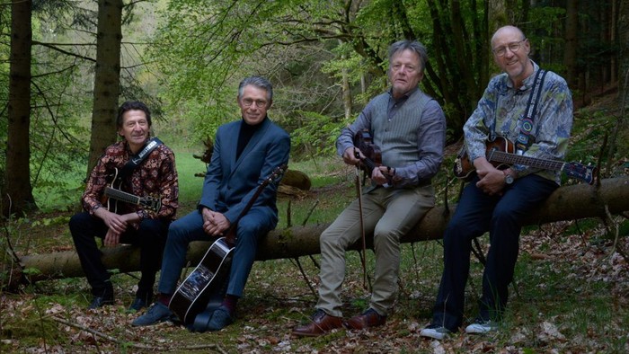 Fire musikere sidder på træstammer i en skov, hver spiller på eller holder guitarer.