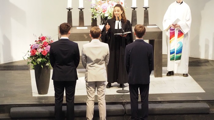 Vier junge Personen und ein Geistlicher nehmen an einer feierlichen Kirchenzeremonie mit Blumenschmuck und Kerzen teil.