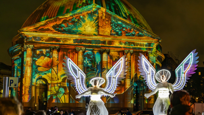 Bunte Lichtprojektionen erleuchten nachts ein prächtiges Gebäude, während in der Nähe engelsgleiche Figuren leuchten.