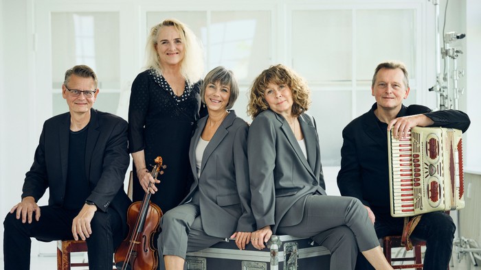 En kvintet af musikere poser sammen med instrumenter, herunder en cello, harmonika og guitarer.