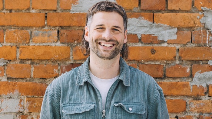 Ein fröhlicher Mann mit langen Haaren lächelt vor einer rustikalen Backsteinmauer, er trägt ein Denim-Hemd über einem weißen T-Shirt.