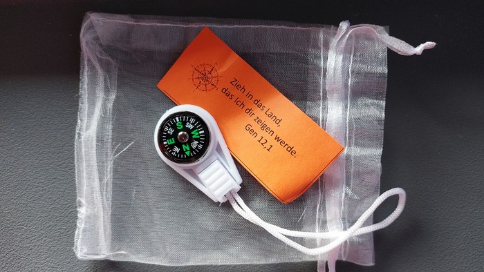 Ein Kompass und ein orangefarbener USB-Stick, eingewickelt in ein Netzbeutel mit deutschem Text.