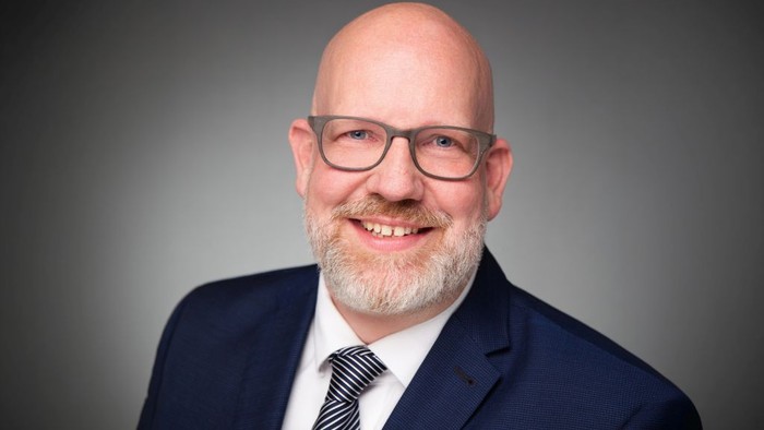 Professioneller Mann im dunklen Anzug, weißem Hemd und gestreifter Krawatte, der in die Kamera lächelt.