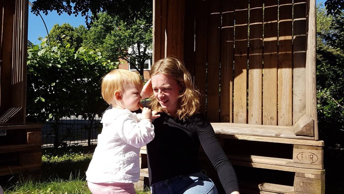 En mor og sit lille barn sitter på en purple dukkeklæde ved en trækonstruktion i en have.