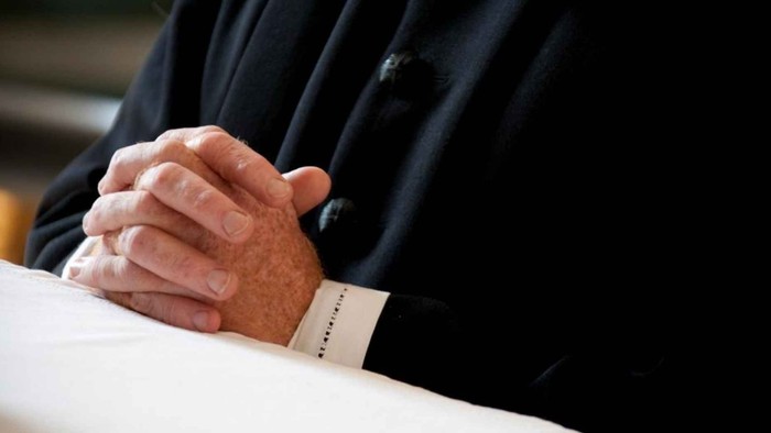 En person i formelt tøj har hænderne foldet sammen i en bønlignende stilling.