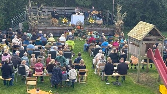 Open-Air-Veranstaltung mit Publikum, das auf Bänken sitzt und eine Bühnenaufführung in einer ländlichen, grasbewachsenen Umgebung verfolgt.