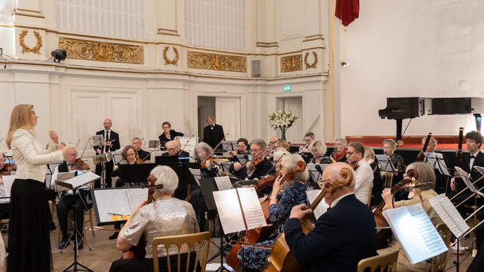 Et orkester optræder i en elegant koncertsal med publikum siddende og en dirigent, der leder musikerne.