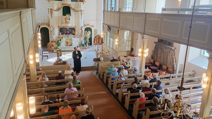 Historisches Kircheninneres mit sitzender Gemeinde während eines Gottesdienstes, mit prunkvollen Altarverzierungen und hölzernen Kirchenbänken.