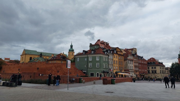 Bunte historische Gebäude umgeben einen lebhaften offenen Platz, auf dem Menschen spazieren, mit einer Backsteinmauer im Hintergrund.