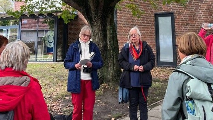 Eine Gruppe von etwa zehn Personen steht im Freien unter einem großen Baum vor einem Backsteingebäude. Zwei Personen stehen in der Mitte der Gruppe, eine davon liest von einem Blatt Papier vor, während die anderen aufmerksam zuhören.