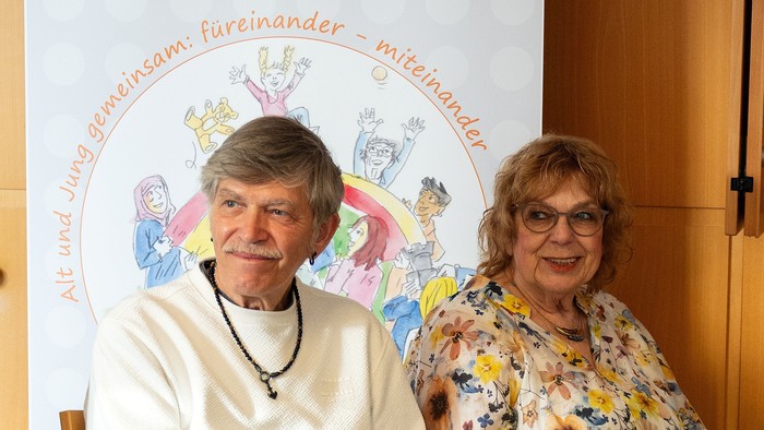 Zwei Menschen sitzen nebeneinander bei einer Veranstaltung. Im Hintergrund steht ein Banner mit einer gezeichneten Gruppe von Menschen unter einem Regenbogen und dem Schriftzug „Alt und Jung gemeinsam: füreinander – miteinander“.