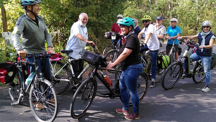Eine Gruppe von Radfahrerinnen und Radfahrern mit Fahrradhelmen steht bei sonnigem Wetter mit ihren Rädern auf einem asphaltierten Weg am Waldrand. Einige unterhalten sich, andere blicken lächelnd in die Kamera.