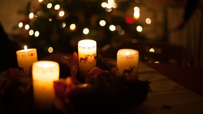 Varmt oplyste stearinlys på et festligt bord pyntet med julegrønt og et sløret juletræ i baggrunden.