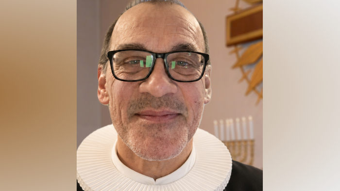 En smilende mand med briller og en hvid rullekravesweater på en neutral baggrund.