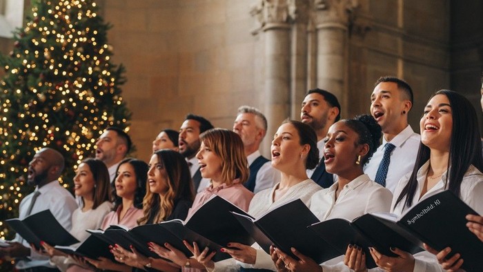 Ein vielfältiger Chor singt leidenschaftlich in einem prächtigen Saal während eines festlichen Weihnachtskonzerts mit einem geschmückten Baum.