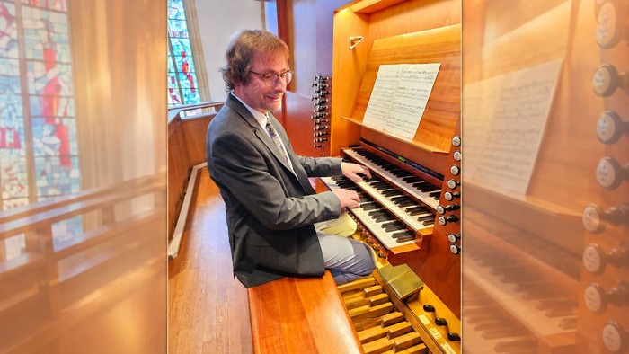 Ein Mann spielt eine Orgel in einer Kirche mit bunten Glasfenstern und ausgelegter Notenmusik.