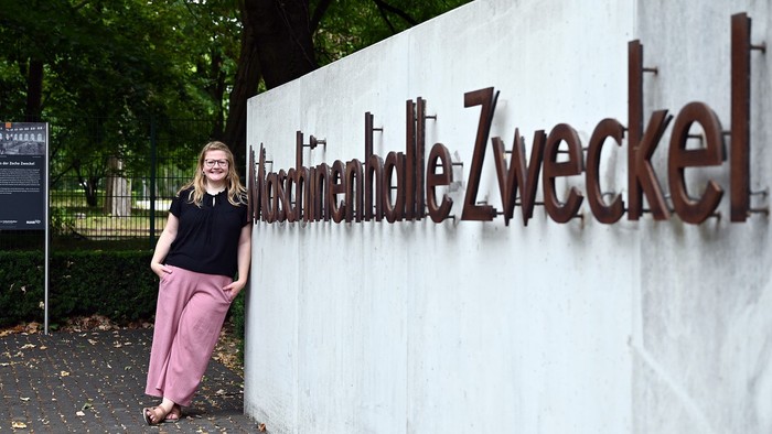 Eine Frau posiert neben einer Mauer mit großem Text „Naturdenkmal Zweckelei“ in einer parkähnlichen Umgebung.