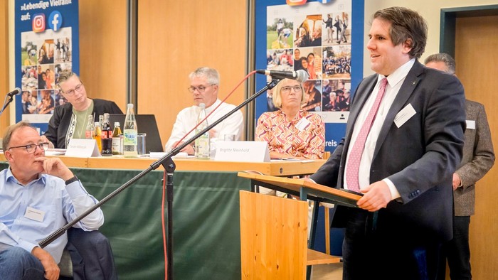 Ein Mann spricht am Mikrofon vor mehreren Zuhörenden