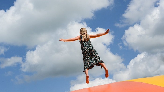 Ein junges Mädchen in einem geblümten Kleid springt mit ausgestreckten Armen auf einer aufblasbaren Spielstruktur vor einem Hintergrund aus blauem Himmel und verstreuten Wolken. 