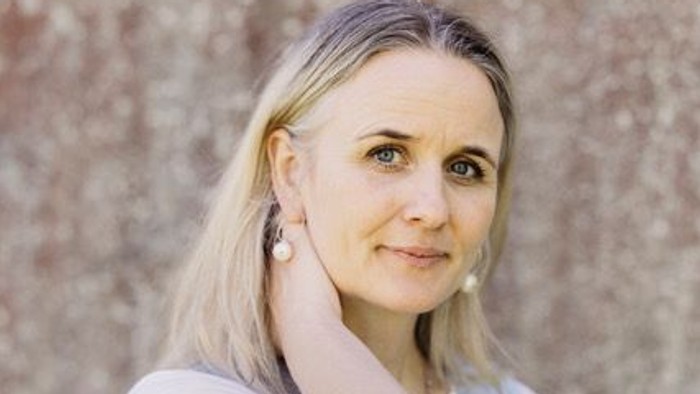 En person med blondt hår og lilla striber, der smiler foran en tekstureret stenmur.