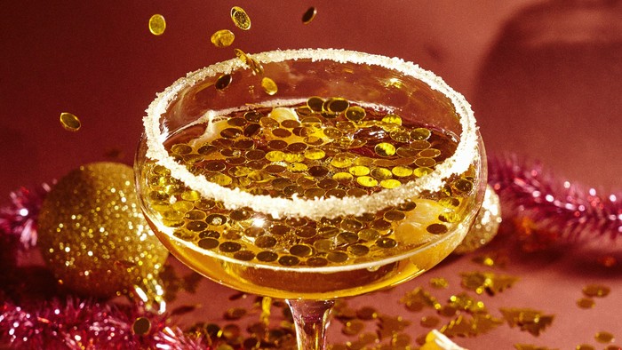 En glashylle med en guldfarvet drink og sukkerrand, omgivet af glitrende dekorationer og guldfarvede smukkelser.