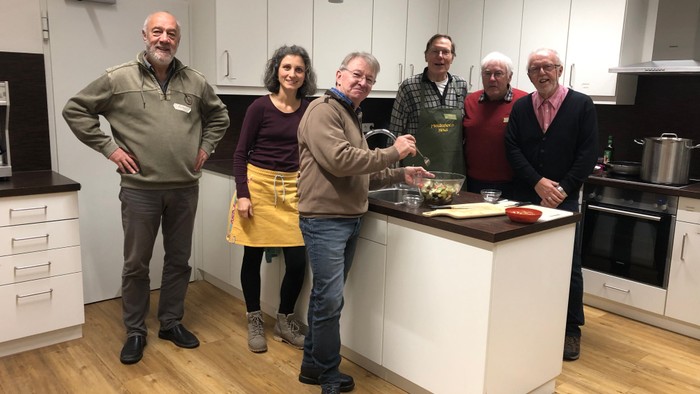Eine Gruppe von sechs Personen, die gemeinsam in einer modernen Küche posieren und Essen zubereiten.
