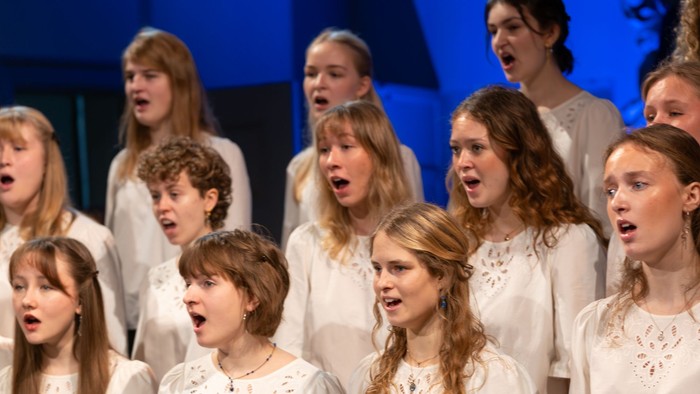 En gruppe unge kvinder i koordinerede hvide toppe og mørke bukser, der synger lidenskabeligt på scenen.