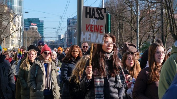 Eine große Menschenmenge protestiert mit Schildern, auf denen „Hass keine Menschen“ steht, in einer städtischen Umgebung.
