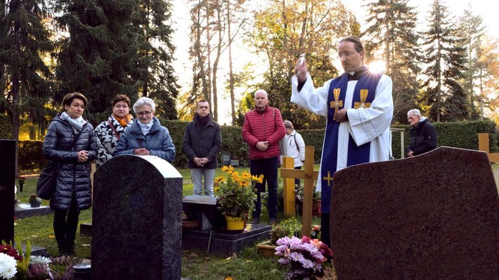 Ein Priester leitet eine Trauerfeier auf einem Friedhof, während Trauernde sich um mit Blumen geschmückte Gräber versammeln.