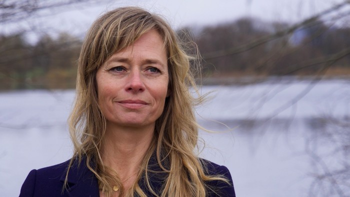 En person med langt blondt hår står udendørs nær et vand, iført en mørk blazer.