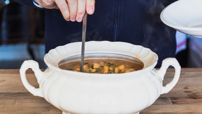 Eine Person serviert Suppe aus einer weißen Schüssel auf einem Holztisch.