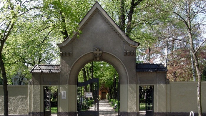 Das Bild zeigt einen Torbogen aus Stein mit einem Kreuz darüber, umgeben von Bäumen und einem Zaun.