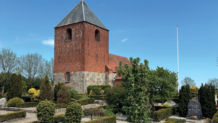 Historisk murstenskirke omgivet af en omhyggeligt anlagt kirkegård med farverige blomster og grønt.