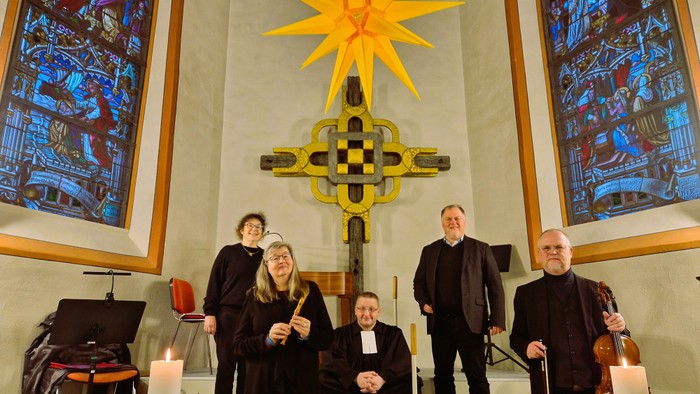 Eine Gruppe von Musikern und Geistlichen posiert in einer Kirche mit farbenfrohen Buntglasfenstern und einem goldenen Kreuz.