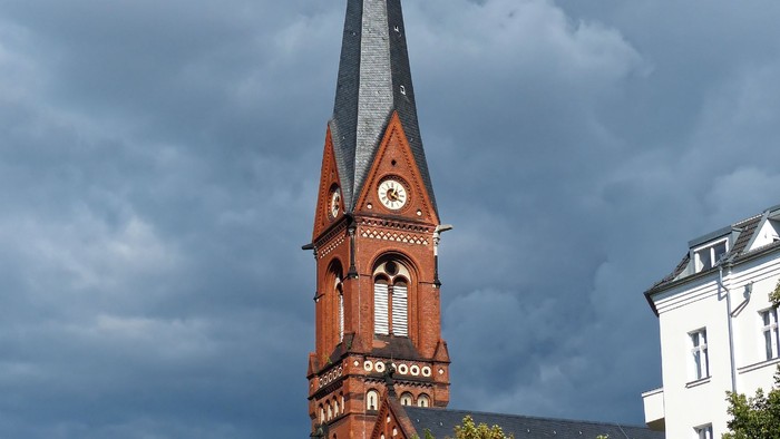 Turm mit Uhr und spitzem Dach, umgeben von Bäumen und Gebäuden