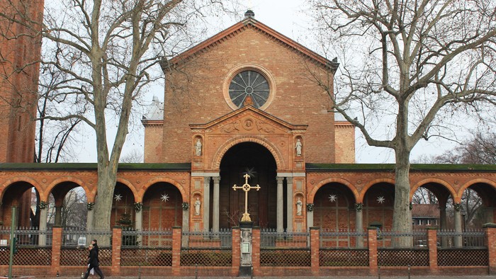 Das Bild zeigt eine Ziegelkirche mit Rundbogenfenstern und einem Kreuz davor.