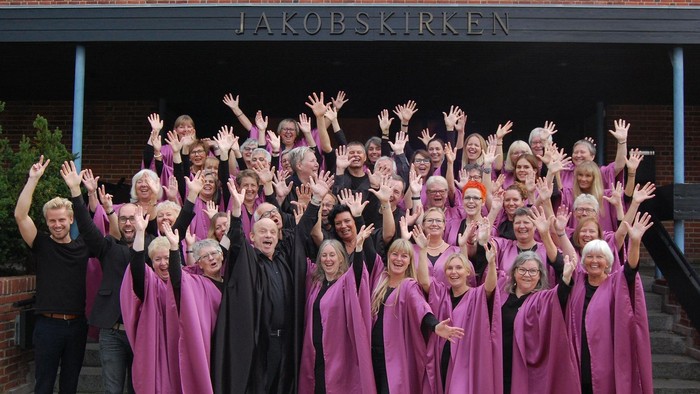 En stor gruppe af kvinder i rosa kapper og sorte skjorter poserer med arme i luften foran Jakobskirken
