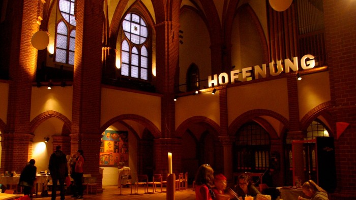 Im Inneren eines großen, gewölbten Raumes sitzen Menschen an Tischen, während ein großes