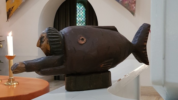 Auf einem weißen Tisch steht eine skurrile Fischfigur vor einem farbenfrohen Gemälde.