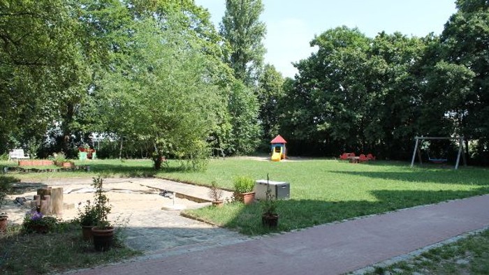Ein ruhiger Parkbereich mit einem kleinen Spielplatz, einer Rasenfläche und schattigen Bäumen.