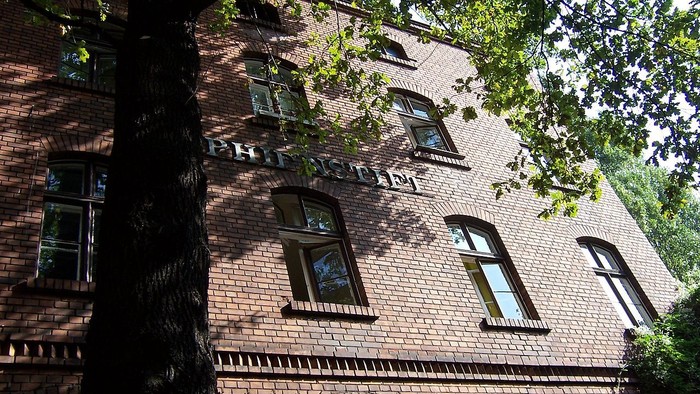 Das Bild zeigt ein rotes Backsteingebäude mit grünen Fensterläden und Bäumen davor.