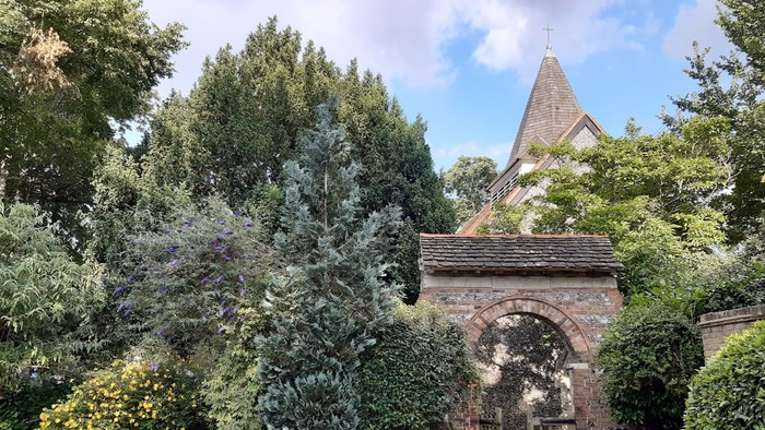 A stone archway surrounded by lush greenery with a pointed roof structure in the background.