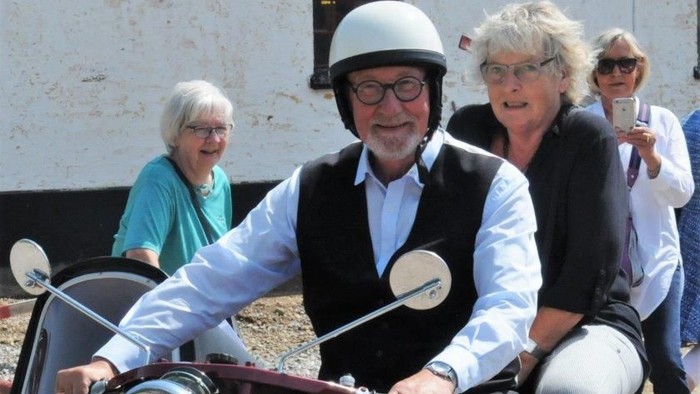 En mand og en kvinde sitter på en rød motorcykel med to andre kvinder i baggrunden.